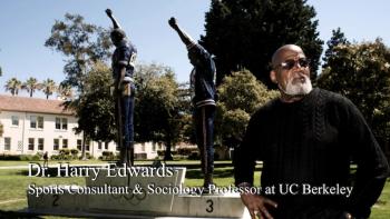 Because They Believed: A man in sunglasses, arms akimbo, stands in front of a statue of Smith and Carlos at the 1968 Olympics
