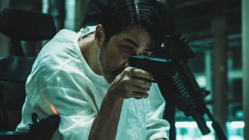 Believer 2 movie: Korean man in white hospital gown center sits holding a gun pointed forward
