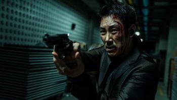Believer movie: Korean man with bloodied face wearing black suit center walks down hall holding up gun pointing left