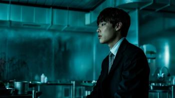 Believer movie: Korean man in black suit, white shirt, and tie on right sits in kitchen with blood sprayed on back wall