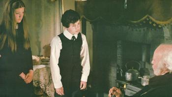The Amazing Mr. Blunden: A young White boy and girl stand in a living space looking down at an old White man as he sits by a fire