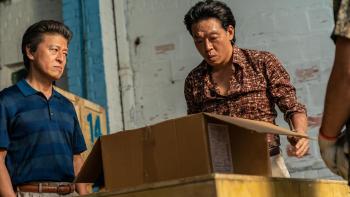 Bogota: City of the Lost movie: Korean man in blue shirt left and Korean man in brown patterned shirt right look in cardboard box in a warehouse