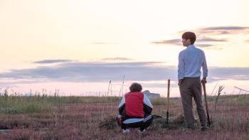 Bogota: City of the Lost movie: In front of recently dug grave, Korean man in tracksuit sits on heels next to Korean man in pants, shirt, and shovel
