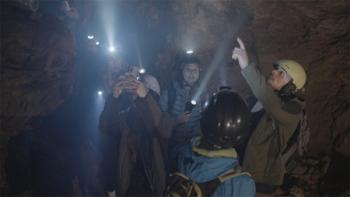 Bone Keeper: A group of people stand inside a cave, which is lit by the beams of their headlamps