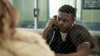 Bosco movie: Black American man center holds a prison phone to his ear and talks to a blond haired visitor with white coat left