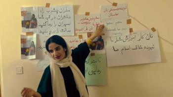 Bread & Roses movie: Afghan woman in white scarf points to the wall behind her that has many pieces of paper with language lessons on them