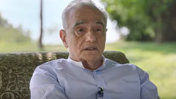 Breakdown: 1975 movie: Martin Scorsese sits on patterned green chair while speaking to the camera, green grass and trees behind him