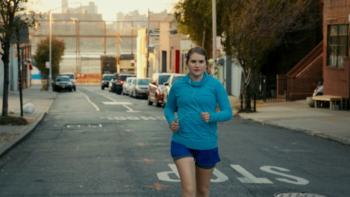 Brittany Runs a Marathon Movie: Brittany runs down the middle of a city street in a turquoise hoodie and blue shorts. 