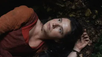 Hamnet: A woman with dark hair lies on the ground among dried leaves and flowers, wearing red and brown period clothing, gazing upward with an emotion