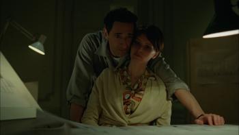 The Brutalist Movie: Erzsébet (Felicity Jones) sits at her husband's desk while he leans into her, his arm around her