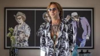 Buen Camino Movie: Man in patterned shirt and sunglasses stands in front of artistic wall portraits.