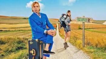 Buen Camino Movie: Man in blue suit sits on suitcase, woman with backpack stands on rural path.