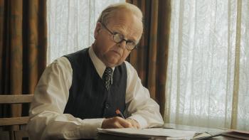 Mr. Burton: Toby Jones sits at a table in front of a window and looks up as he holds a pen