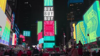 Buy Now! The Shopping Conspiracy poster: A large illuminated sign on the side of a building reads "Buy buy buy"