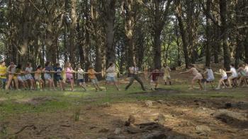Camp Crasher movie: In woodsy area, preteens play tug-of-war, holding onto a long rope either side of a Spanish woman in middle