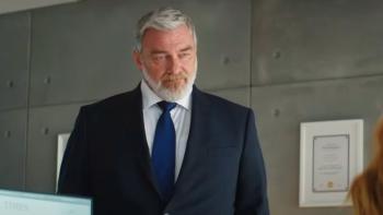 Canary Black: Ray Stevenson, wearing a black suit and blue tie, stands in an office looking down