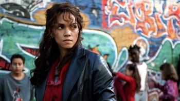 Catwoman movie: Woman in a leather jacket stands in front of a colorful graffiti wall and people in the background
