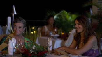Christmas in the Caribbean: Caroline Quentin and Elizabeth Hurley sit at an outdoor dining table, smiling and reaching for each other's hand. 