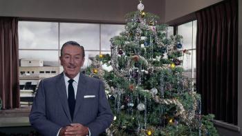 Still from Christmas with Walt Disney: In the background to the right is a decorated Christmas tree, and in the foreground to the left stands award-winning animator Walt Disney who faces the camera to speak about the holiday season, wearing a white shirt, black tie, and blue suit