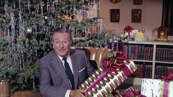Still from Christmas with Walt Disney: In the background to the left is a decorated Christmas tree and to the right a filled bookcase and wall with plaques, and in the foreground Walt Disney is seated at a desk holding a wrapped holiday gift, wearing a white shirt, black tie, and gray suit