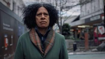 Cousins Movie: An adult Māori woman gazes unhappily from a retail street, wearing a scarf and coat