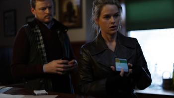 Cult Killer Movie: Private eye Cassie Holt (Alice Eve, right) holds a cell phone and appears to be questioning someone off-camera, while officer Rory McMahon (Paul Reid) stands behind her