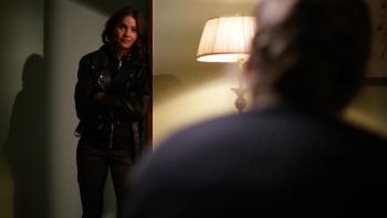 Cult Killer Movie: Revenge-driven Jamie Douglas (Shelley Hennig) leans against a wall in Cassie's apartment, speaking to Cassie, who we can see out of focus and from the back