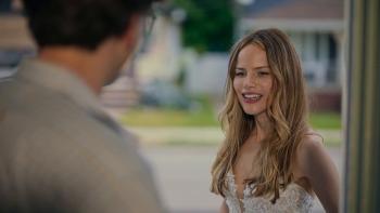 Daughter of the Bride Movie: A young woman from mid length in a bridal gown smiles awkwardly at a blurry male figure with his back to the camera.