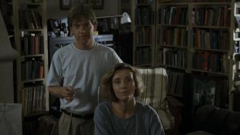 Dead Again: Emma Thompson sits in a chair in a dimly lit living room and Kenneth Branagh stands behind her holding a business card