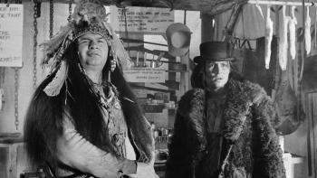 Dead Man: A Native American wearing a headdress and a White man wearing a fur and a black cowboy hat, stand next to each other inside a tent