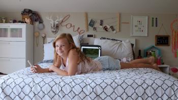 Dear Thirteen movie: Evie lying on bed, smiles and holds a smartphone, wearing white sleeveless top and blue shorts