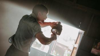 Death Whisperer 2 movie: Indonesian man in blue shirt douses wrapped fist with holy water from a metal flask, light streams in through open window