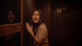 Death Whisperer 2 movie: Indonesian woman with long black hair center inches along hallway wall yelling, her reflection on left in glass window