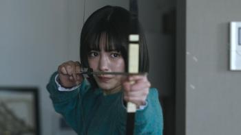 Demon City movie: Japanese teenage girl in teal sweater center draws an arrow with a bow and points it at off-screen target while in a living room