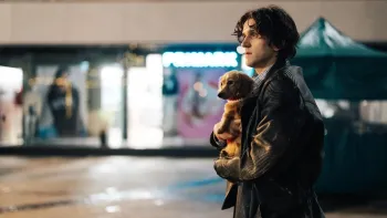 Pillion: A young White man, holding a dog, stares at someone out of shot