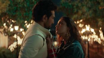Dhoom Dhaam movie: Indian woman in jean jacket right grabs cuff of white suit of Indian man center left, sparkly lights on trees behind them