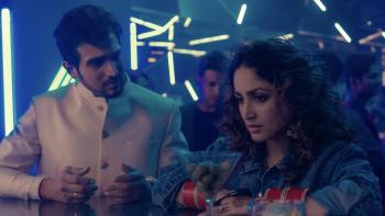Dhoom Dhaam movie: Indian man in white suit at bar in nightclub with Indian woman in jean jacket right, martini glass full of olives in foreground
