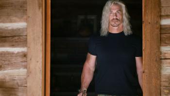 Die Hart 2: Die Harter movie: John Cena in black t-shirt and long blond hair stands under wooden cabin doorway
