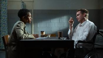 DogMan Movie: Seated at a small table, Evelyn (Jojo T. Gibbs, left), questions Douglas (Caleb Landry Jones), who's smoking