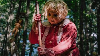 Dolly Movie: A woman in a pink dress and creepy doll mask holds a shovel; dolls are hanging on trees behind her