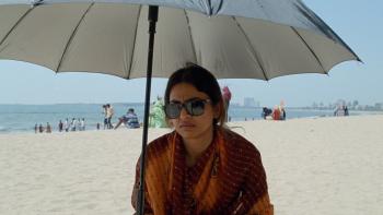 Sister Midnight: An Indian woman wearing sunglasses sits under an umbrella on the beach looking sad