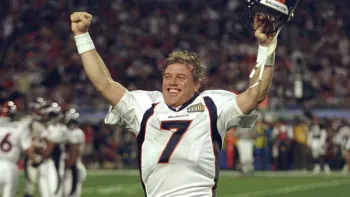 Elway movie: John Elway raises both arms high in the air, with helmet in one hand, in celebration, crowd, players, and stadium in background