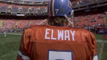 Elway movie: The back of John Elway in Denver Broncos uniform and helmet as he walks on the field, crowd and stadium in the background