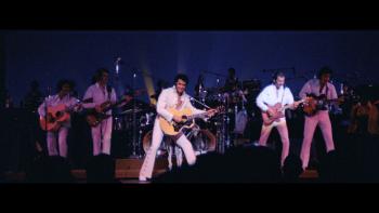 EPiC: Elvis Presley in Concert movie: Presley performs with a wink and a smile, holding his guitar to the right with his band members similarly posed