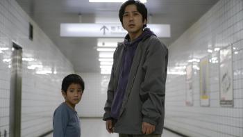 Exit 8 Movie: A young boy and the lost man stand in a subway corridor looking at something as if considering it