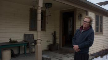 Father Mother Sister Brother movie: Older man with glasses stands on a porch outside a rural home, wearing a blue jacket and burgundy scarf