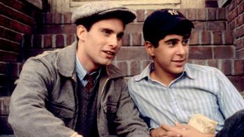 Two young men with dark hair lounge outdoors, one wears a paperboy cap