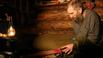 Happy People: A Year in the Taiga Movie: Thin White man in his 50s with beard and gray sweater sits inside a rustic cabin