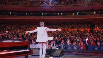Fito Páez in white facing cheering audience in a large theater.