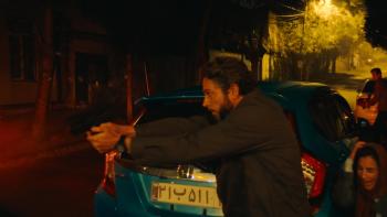 The Fix Movie: In Tehran, Tucker (Zachary Levi) points a gun while a scared woman crouches behind a car